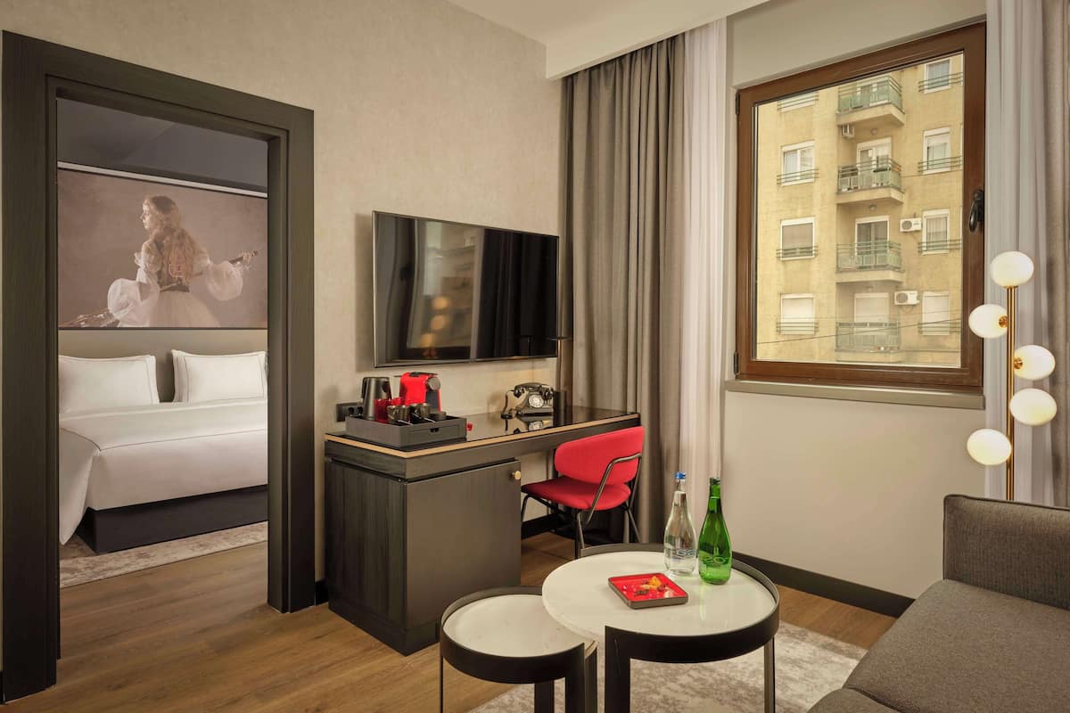 Une chambre d’hôtel avec un lit, un bureau avec une télévision, une chaise rouge, une petite table avec une bouteille, et une fenêtre donnant sur un immeuble.