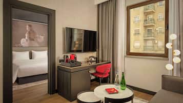 Une chambre d’hôtel avec un lit, un bureau avec une télévision, une chaise rouge, une petite table avec une bouteille, et une fenêtre donnant sur un immeuble.