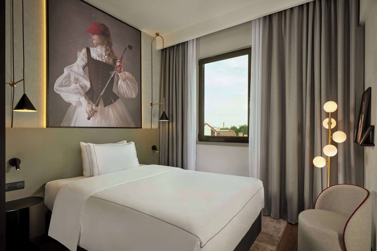 Une chambre d’hôtel avec un grand lit, une table de chevet, une chaise, une grande fenêtre avec des rideaux et un tableau accroché au mur.