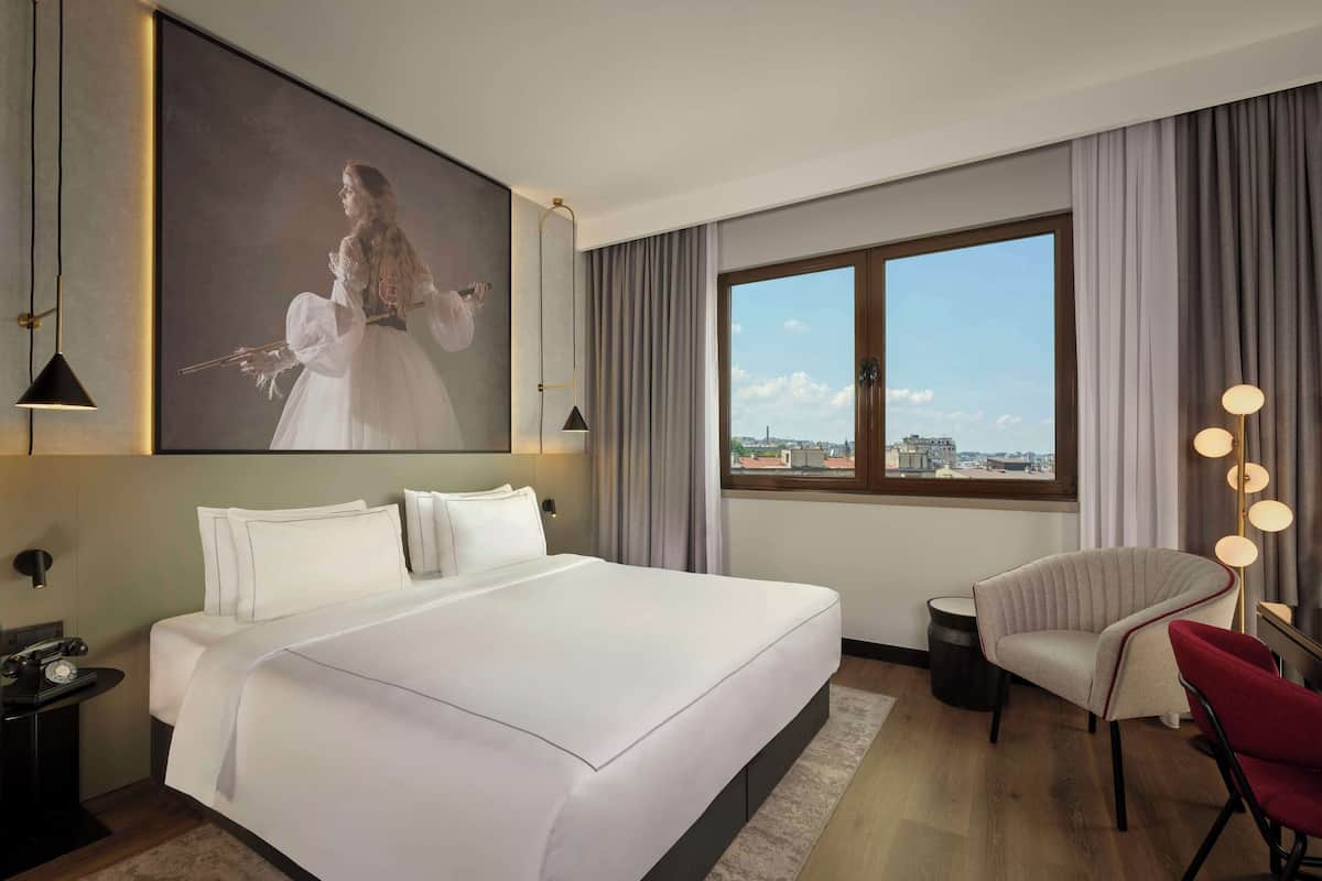 Une chambre d’hôtel avec un grand lit, un bureau avec une lampe, une chaise, une grande fenêtre donnant sur la ville et un tableau au mur.