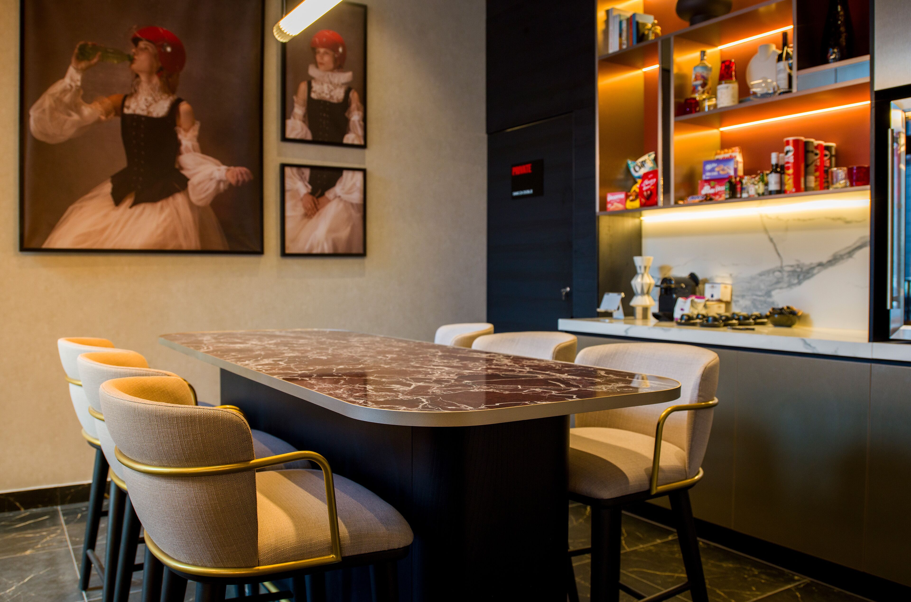 Un espace bar moderne avec une table surmontée d’un plateau en marbre, des chaises rembourrées et une étagère bien garnie de différentes bouteilles.