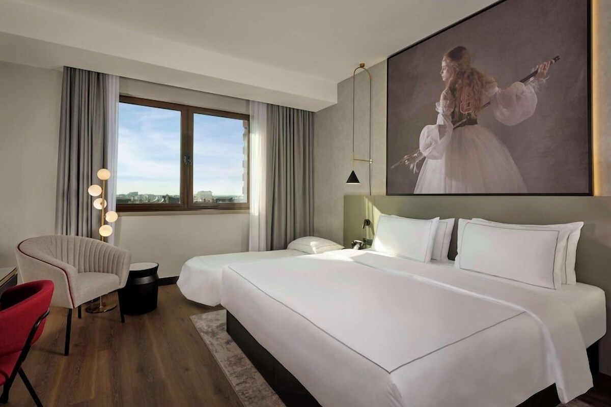 A hotel room with a large bed, a desk, a chair, and a large window.