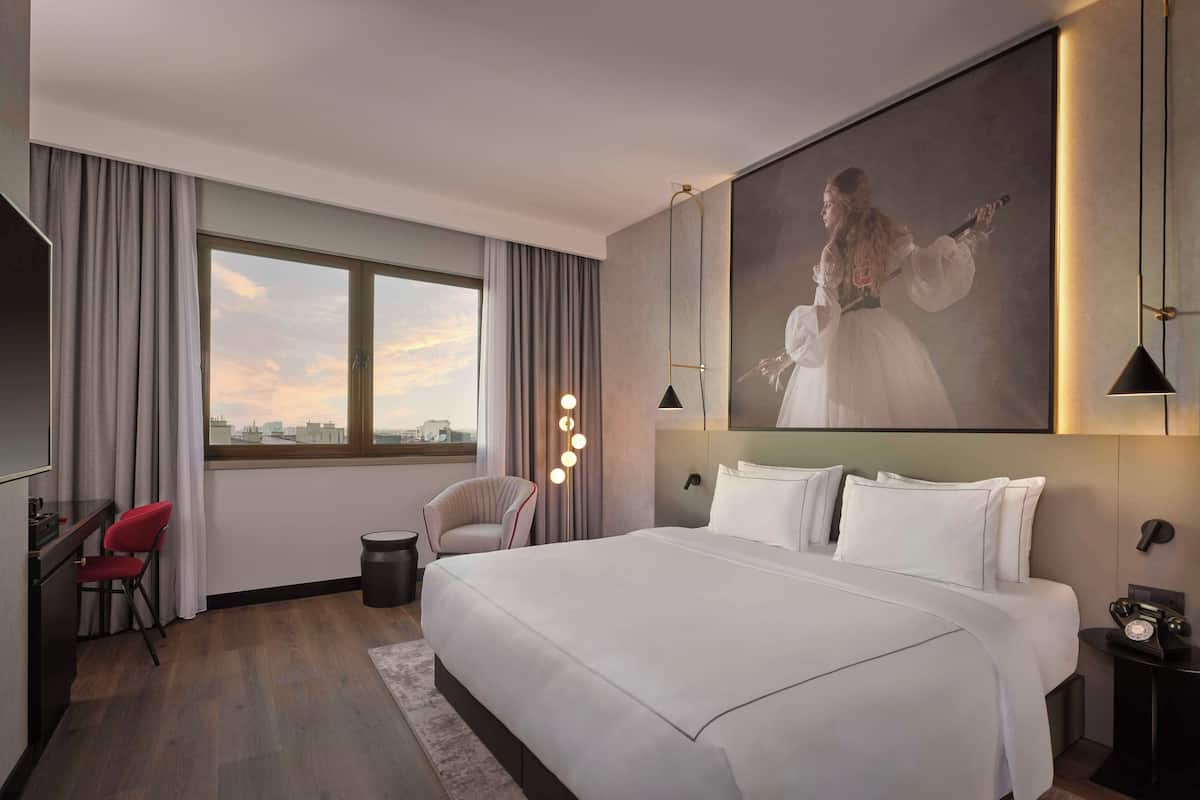 Une chambre d’hôtel moderne, dotée d’un grand lit, d’un bureau, d’une chaise et offrant une vue sur la ville grâce à une grande fenêtre.