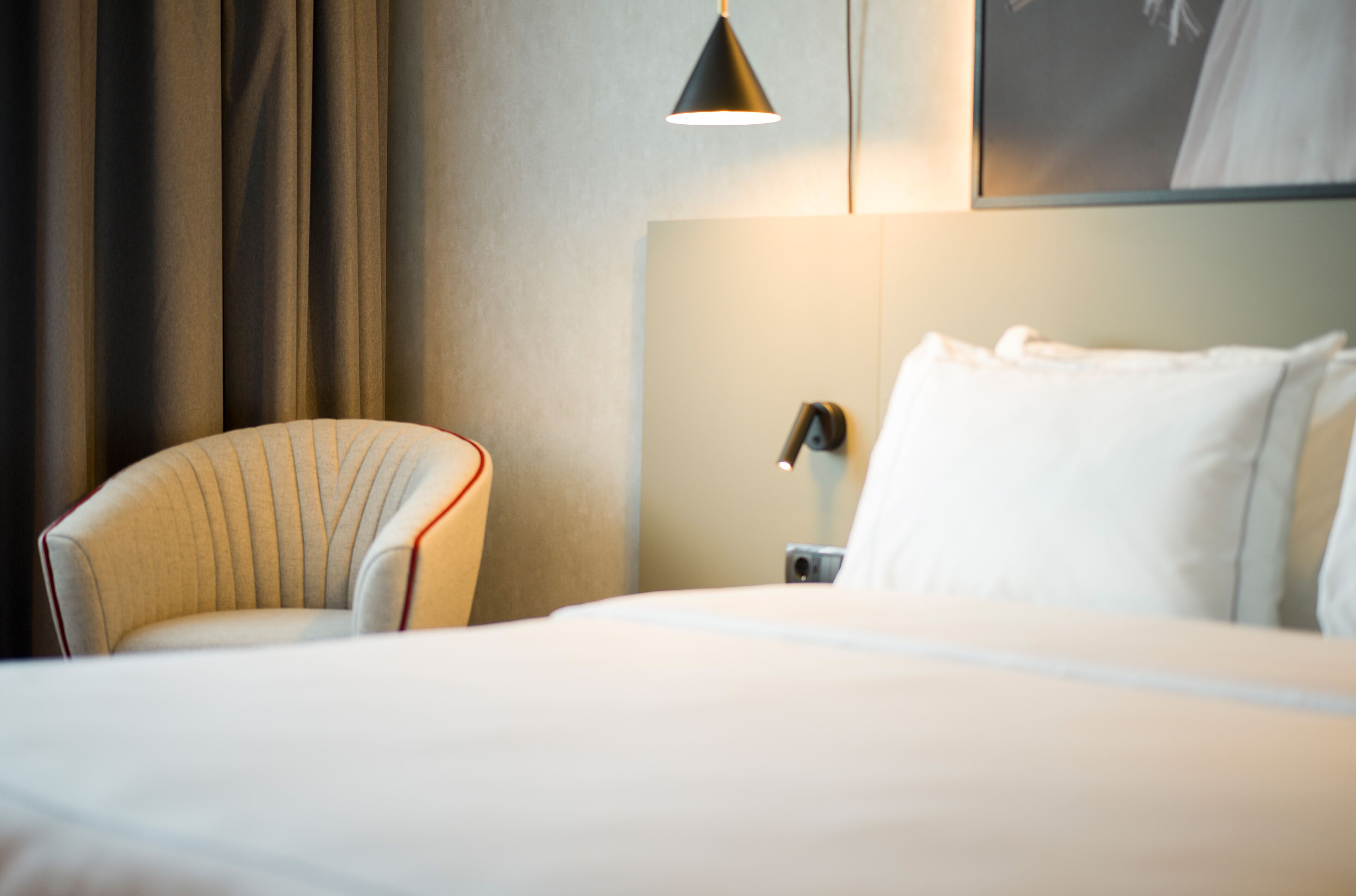 A hotel room with a bed, a chair, a nightstand, a lamp, and curtains.