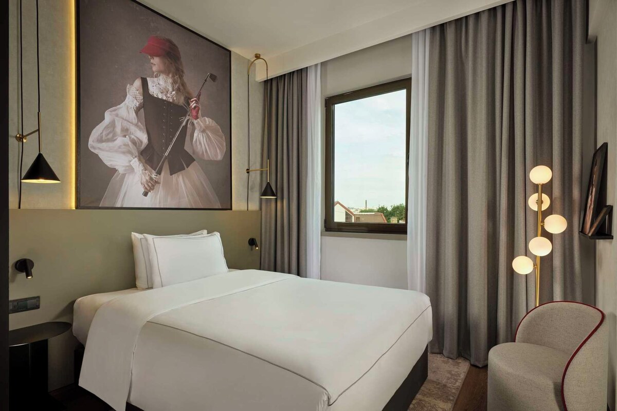 A hotel room with a large bed, a nightstand, a desk, a chair, a window with curtains, and a wall-mounted lamp.