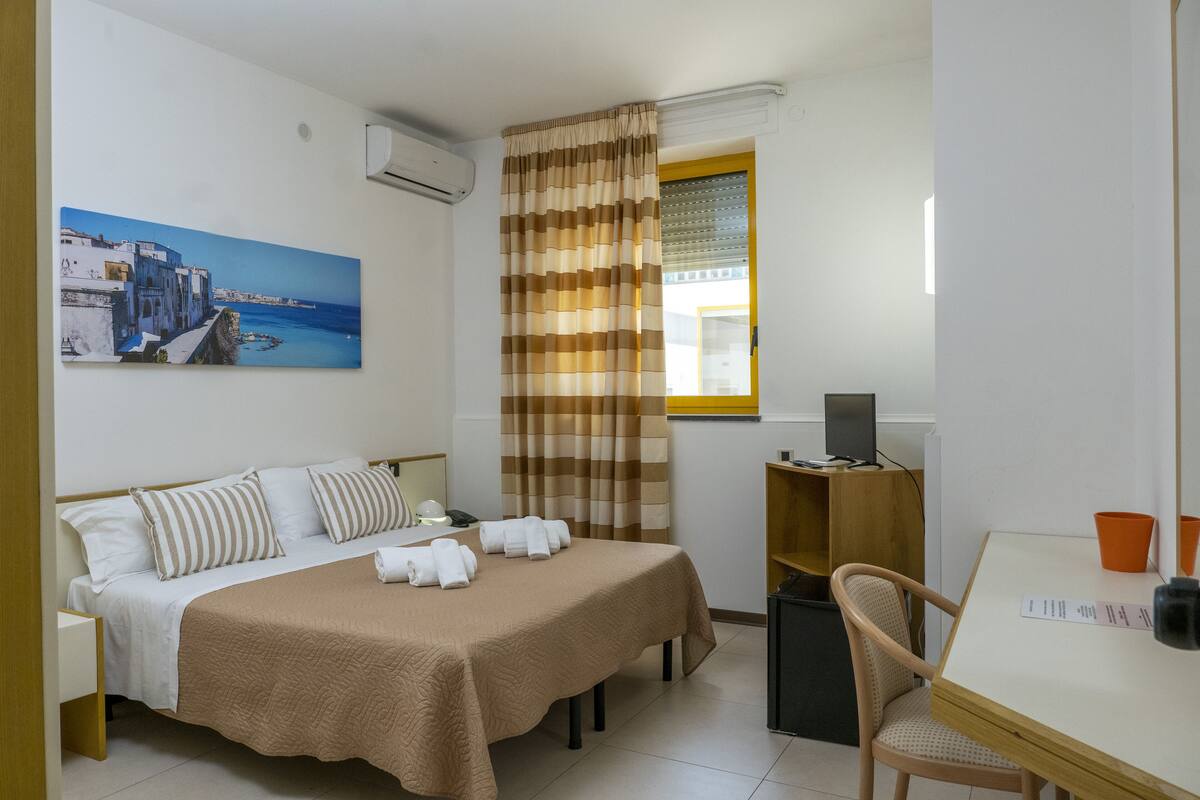 A hotel room with a bed, a desk, a chair, a window with curtains, a painting on the wall, and a TV on a stand.