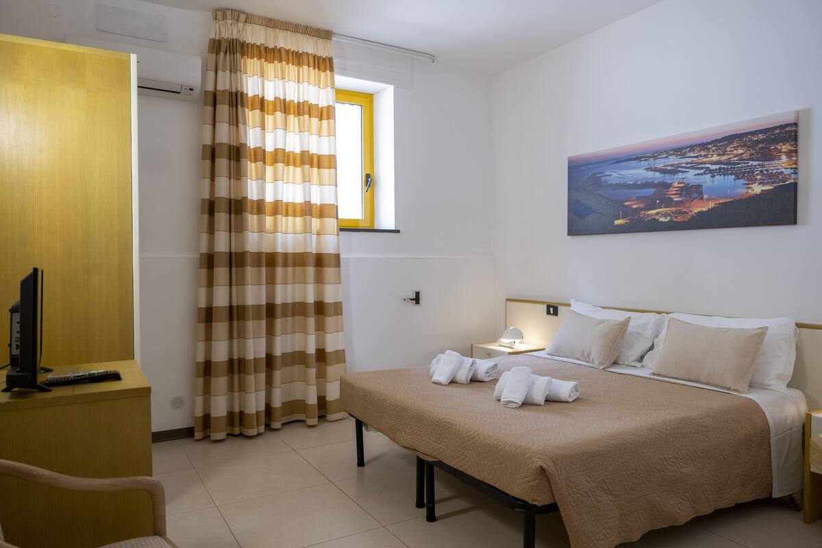 A hotel room with a bed, a desk with a computer, a window with curtains, and a painting on the wall.