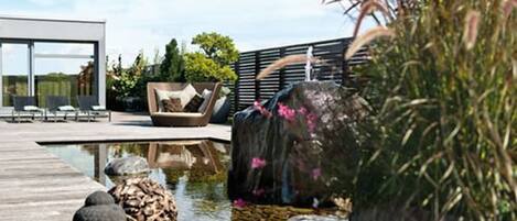 A modern outdoor pool area with a wooden deck, a seating area with cushions, and a waterfall feature.