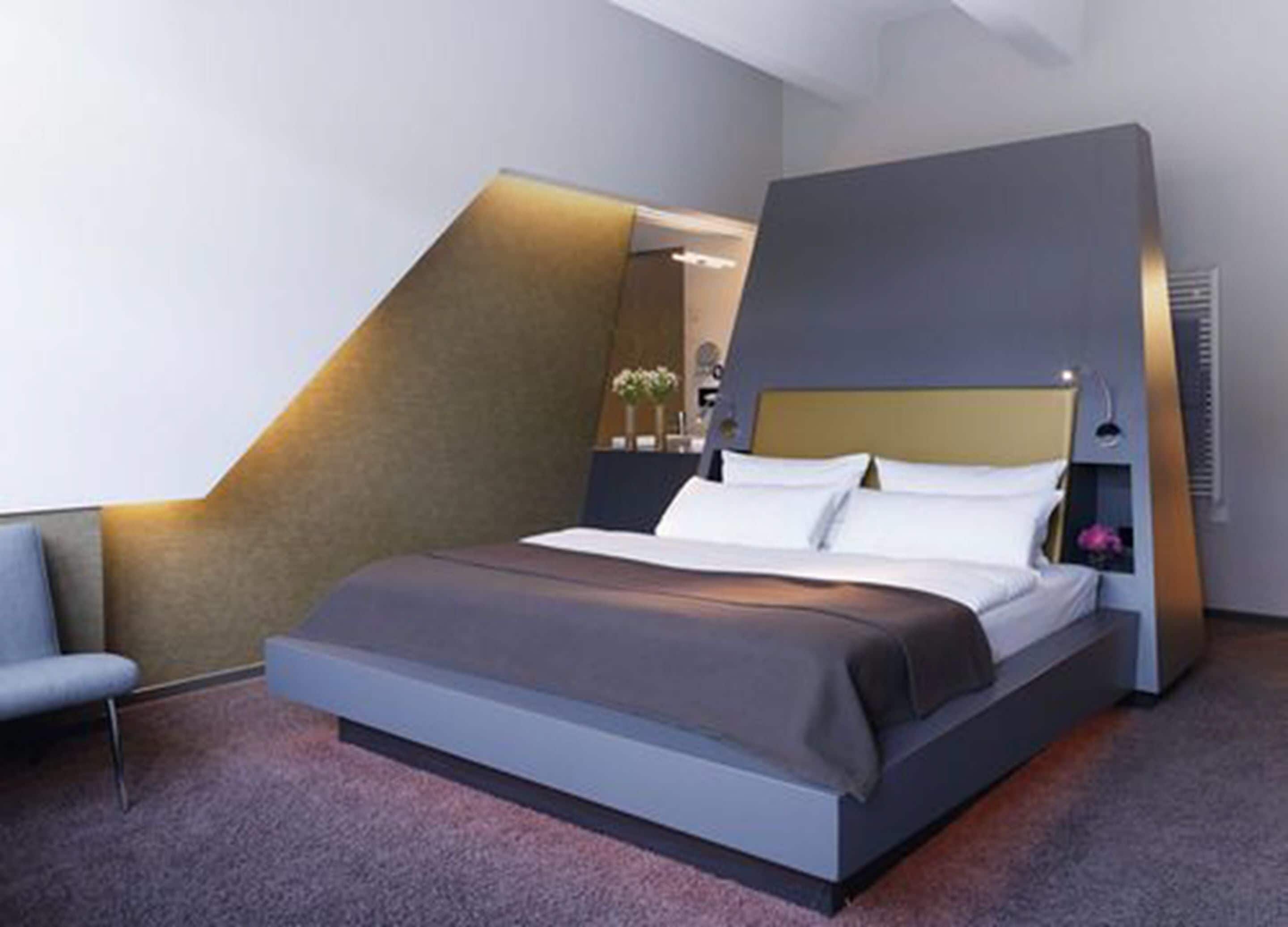 A modern bedroom with a large bed, a chair, and a unique sloped ceiling design.