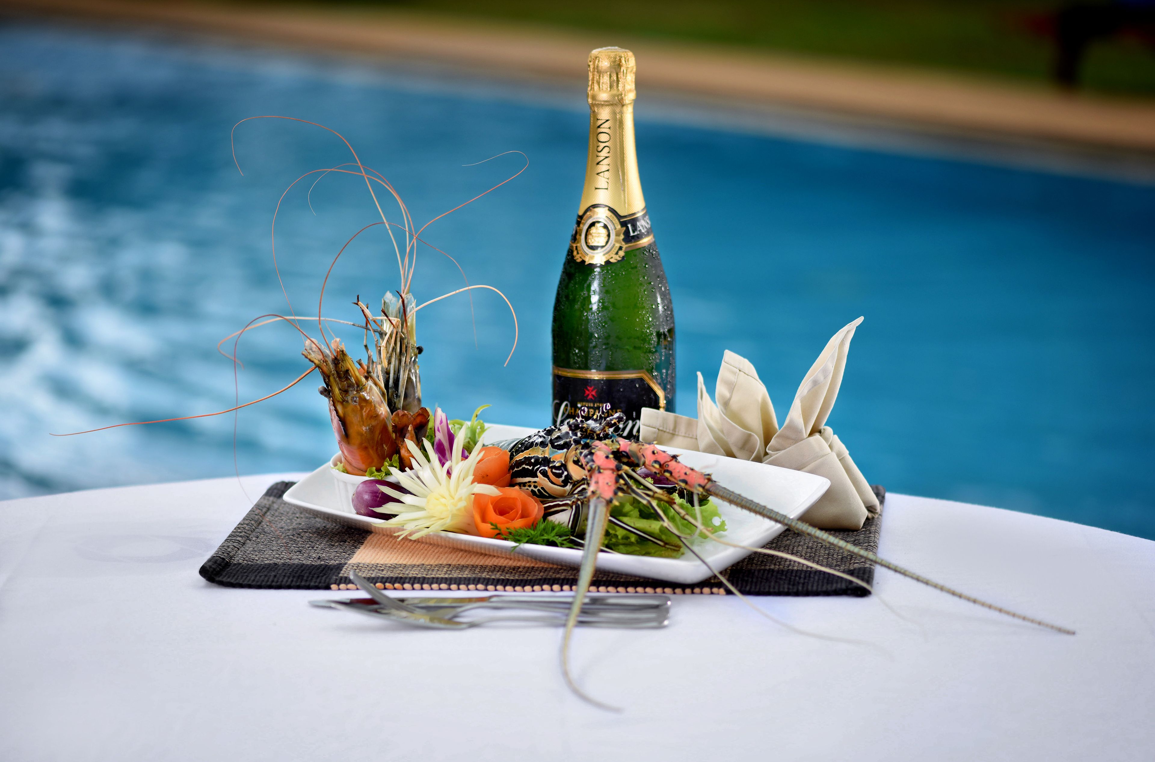 A table with a bottle of champagne, a lobster, and a plate of vegetables by a pool.