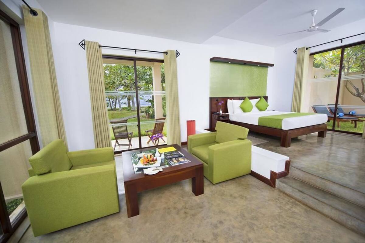 A hotel room with a bed, two green armchairs, a coffee table, and a large window with curtains.
