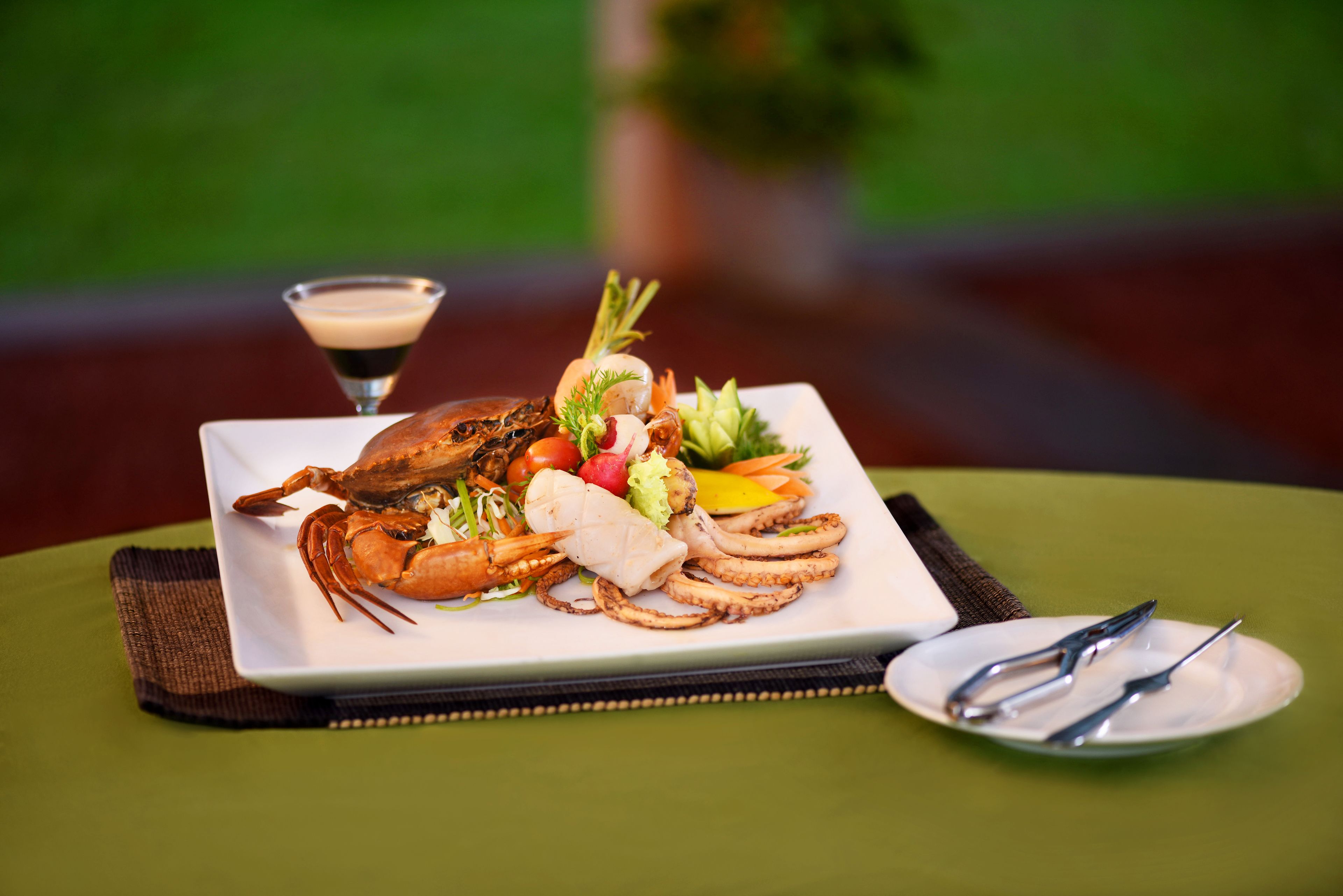Una bandeja de mariscos con langosta, camarones y pulpo, sobre un plato blanco, acompañada de una copa de martini y cubiertos sobre un mantel verde.