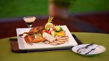 Una bandeja de mariscos con langosta, camarones y pulpo, sobre un plato blanco, acompañada de una copa de martini y cubiertos sobre un mantel verde.