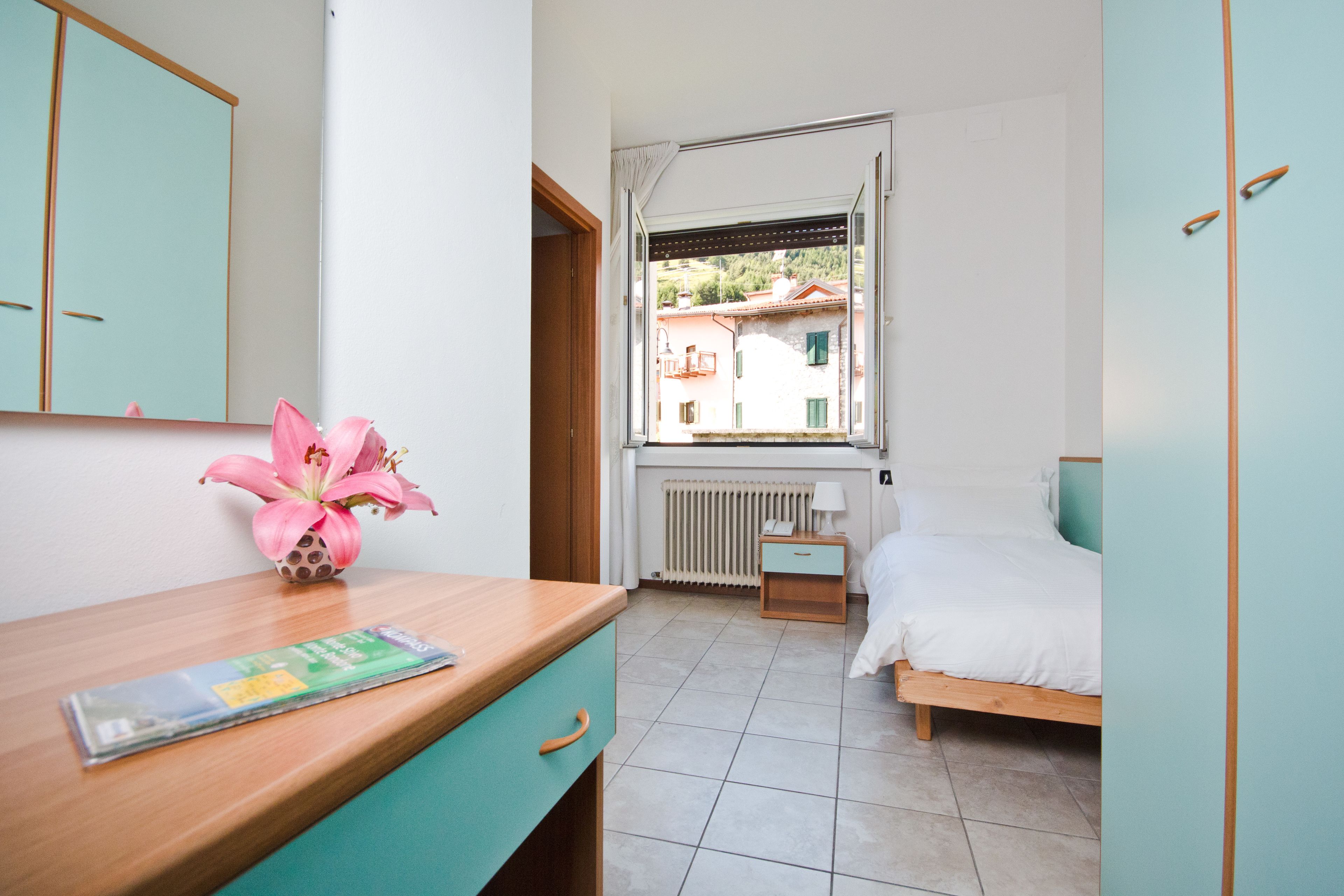 Un dormitorio con un escritorio de madera, un jarrón de flores rosadas y una cama con ropa de cama blanca, con vistas a edificios a través de una ventana abierta.