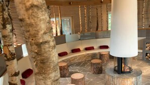 Un espace de détente circulaire en pierre, avec un foyer central, entouré de tabourets en bois et de coussins rouges.