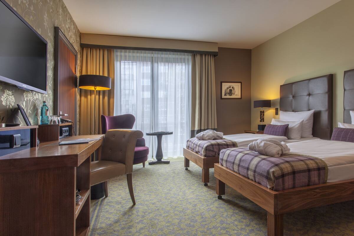 Habitación de hotel con cama, dos mesitas de noche, una silla, escritorio, televisión y ventana con cortinas.
