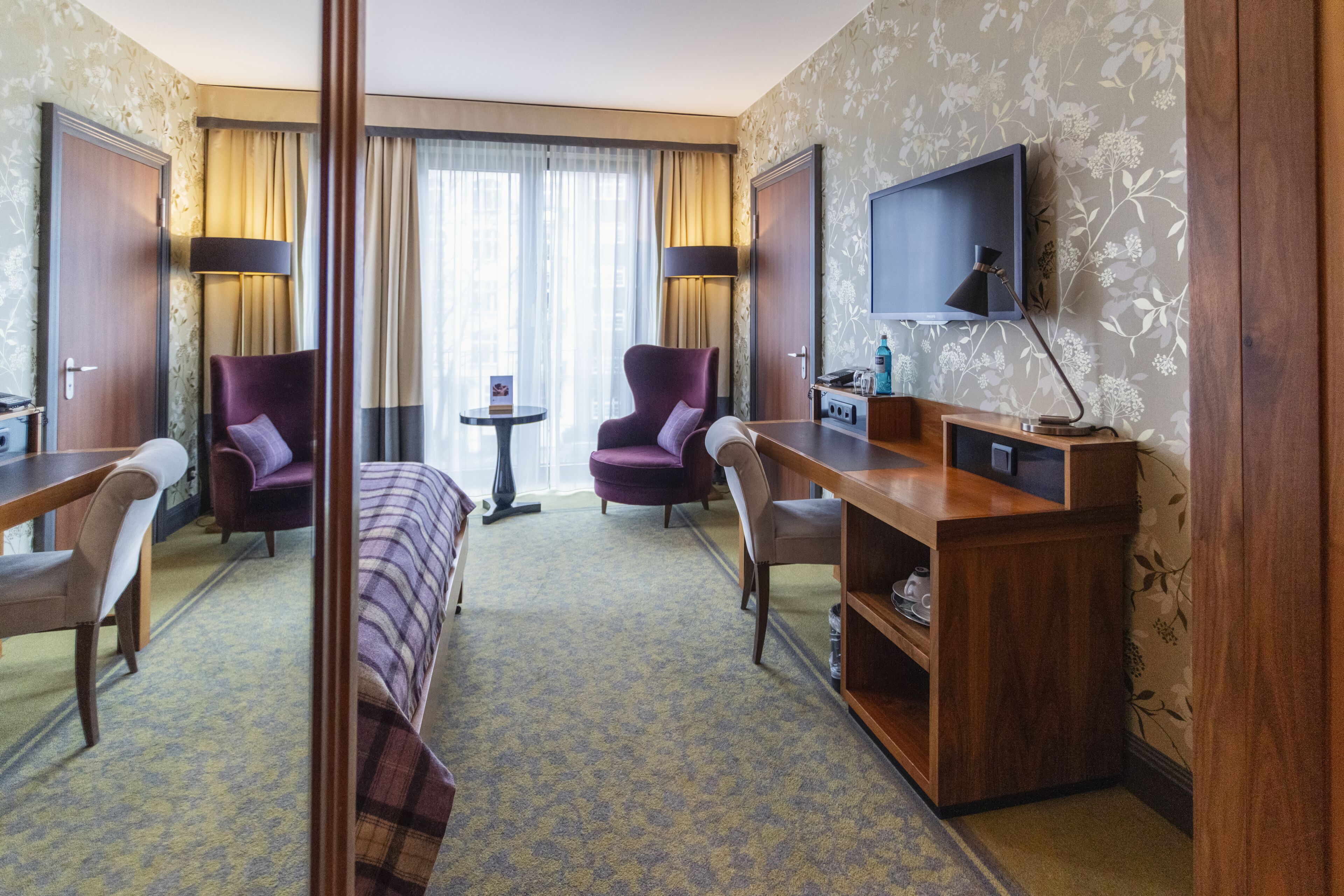 A hotel room with a bed, two armchairs, a desk, a TV, and a small table with a lamp.