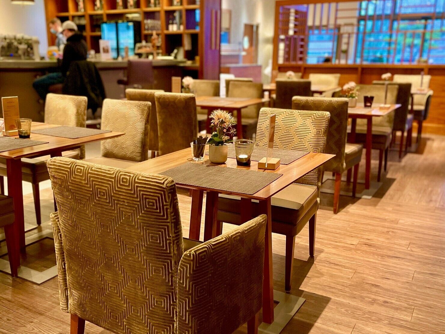 Ein Restaurant mit Holztischen und -stühlen, einer Bar und Blick auf die Stadt durch große Fenster.