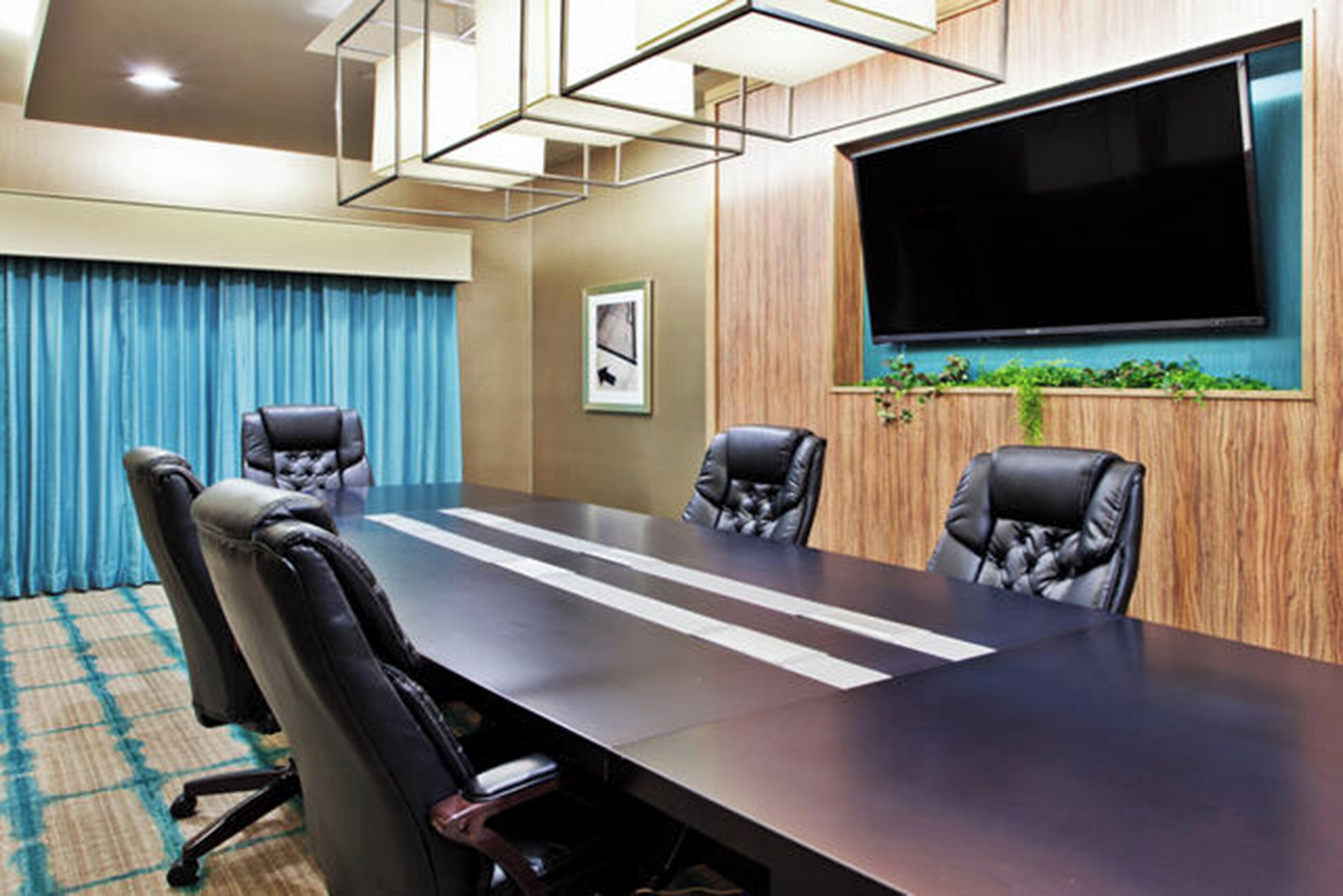 A conference room with a long table, four chairs, a large flat-screen TV, and a framed picture on the wall.
