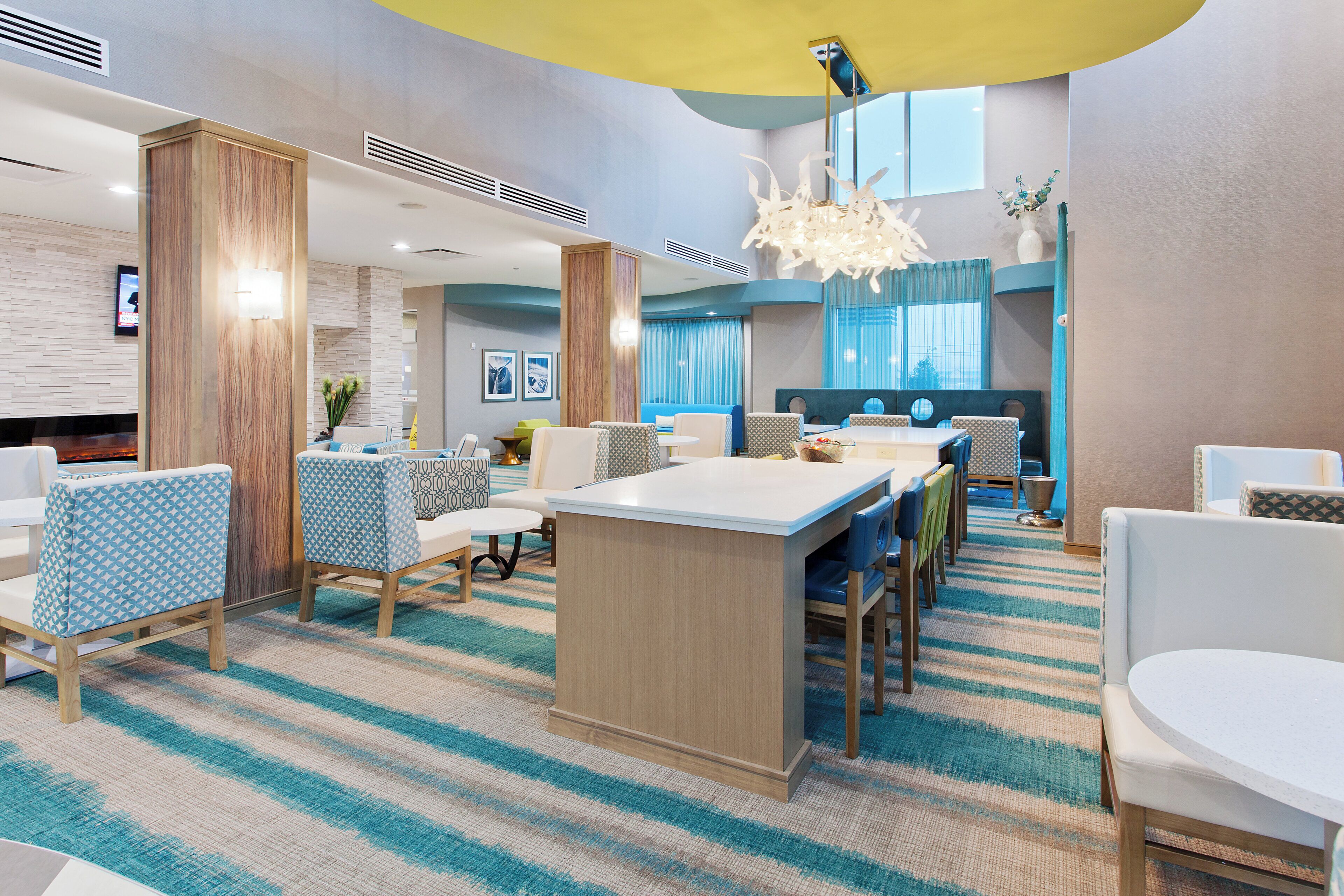 Eine moderne Hotellobby mit einer zentralen Insellounge, einer Sitzecke mit gemusterten Stühlen und einem großen Fenster mit Vorhängen.