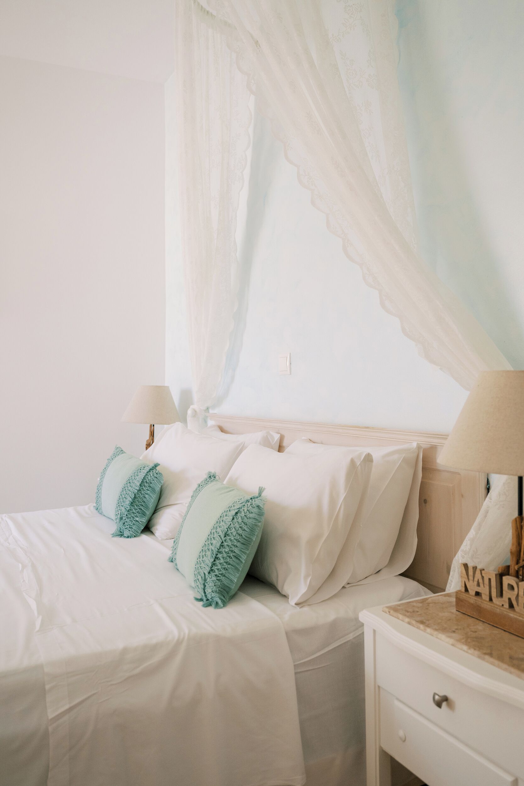 A neatly made bed with white bedding and teal pillows, a bedside table with a lamp, and a wall-mounted nightstand.
