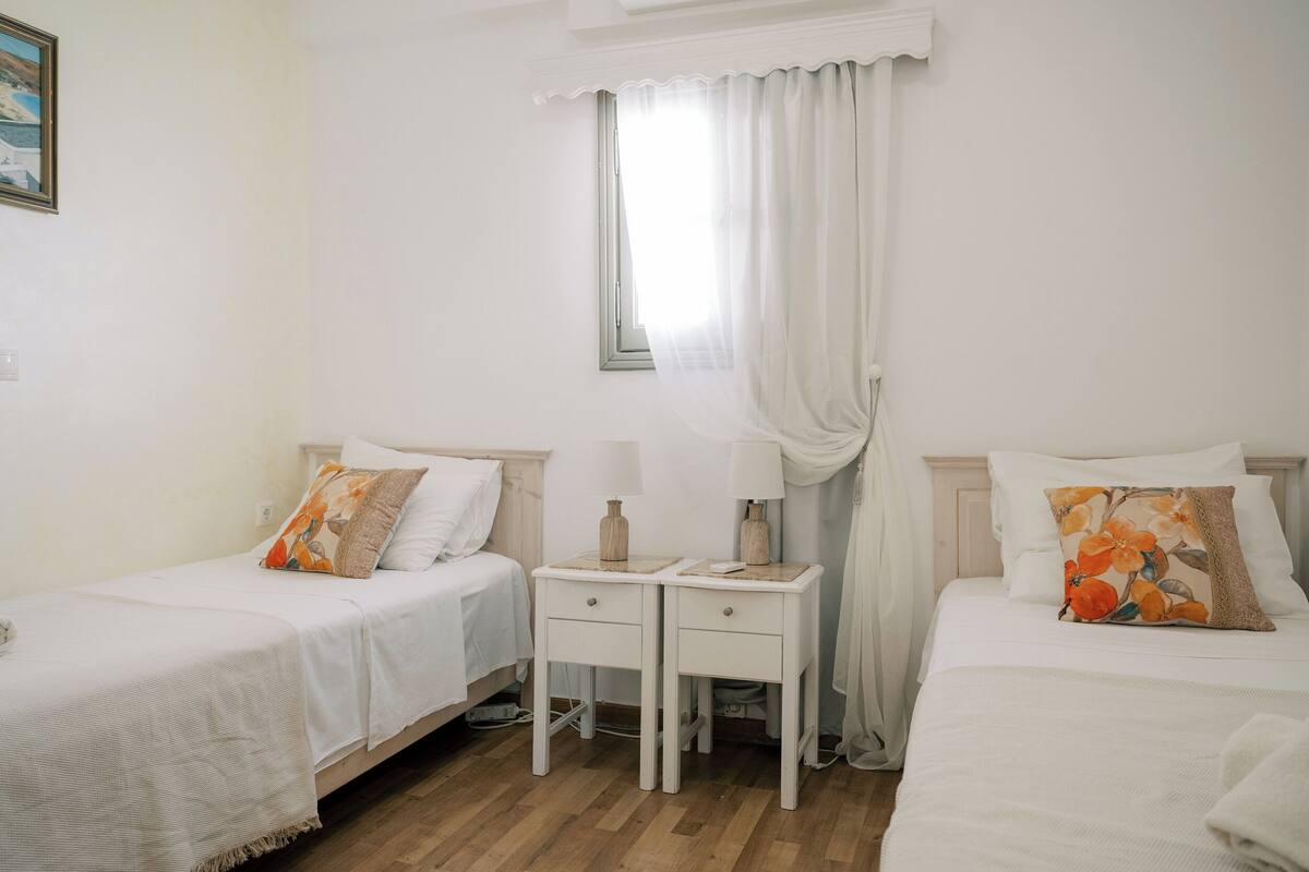 Un dormitorio con dos camas individuales, una mesita de noche con lámpara y una ventana con cortinas.