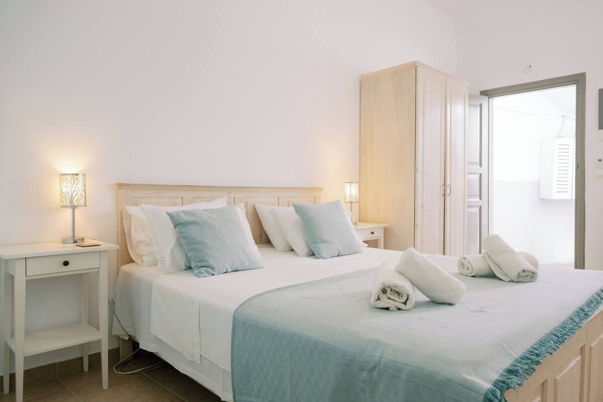 Una cama bien hecha con ropa de cama blanca y azul claro, dos mesitas de noche con lámparas y un armario de madera.