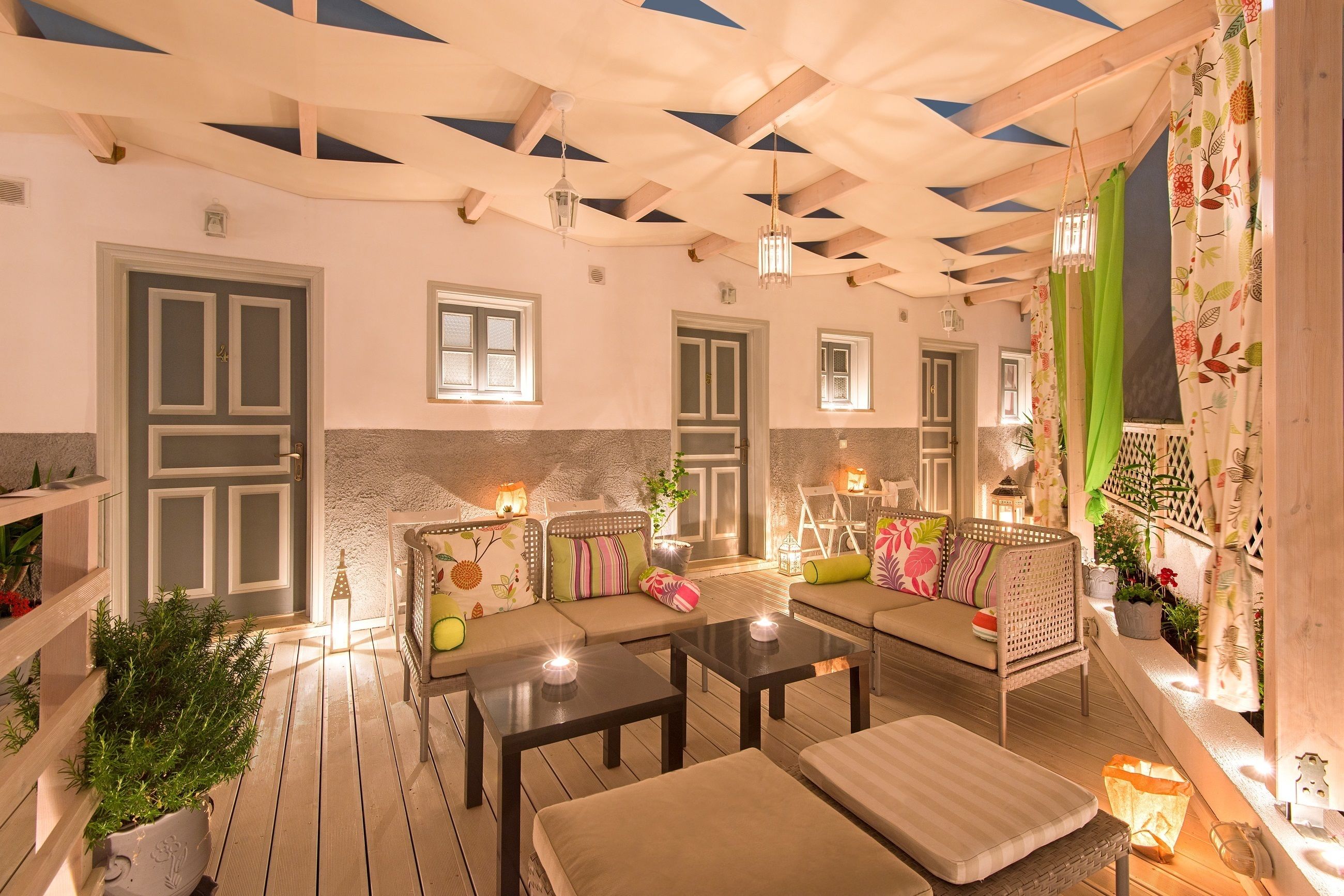 Un rincón al aire libre acogedor, con mesas de madera y sillas con cojines, rodeado de plantas en macetas e iluminación decorativa.