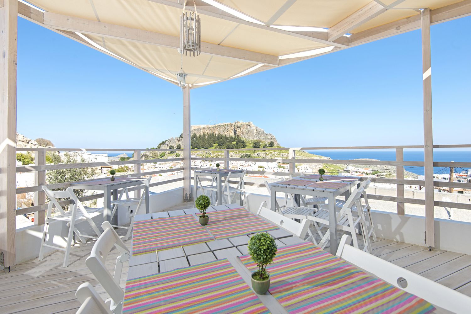 Un patio al aire libre con mobiliario blanco y manteles a rayas, con vistas a la costa y una colina de fondo.