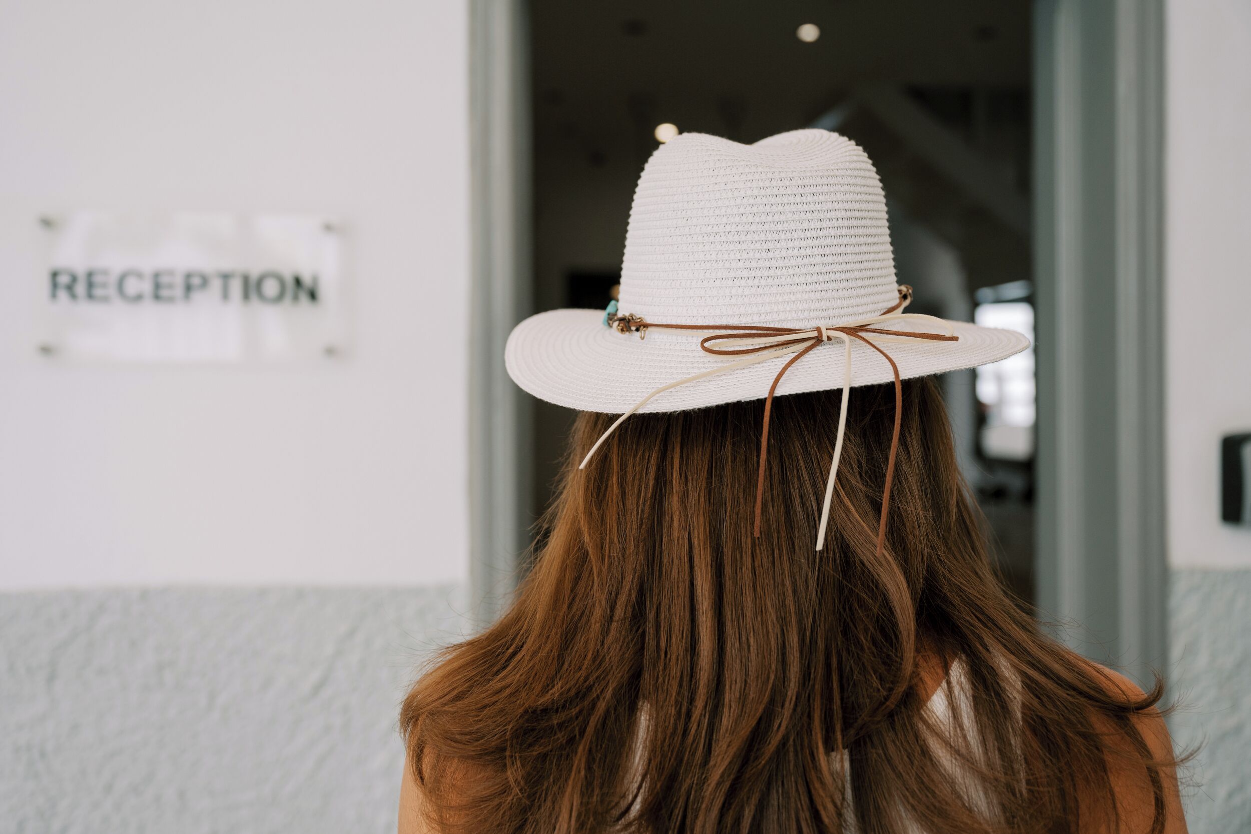 Una persona con capelli lunghi indossa un cappello bianco con un nastro marrone, in piedi di fronte a un cartello con la scritta 'RECEPTION'.