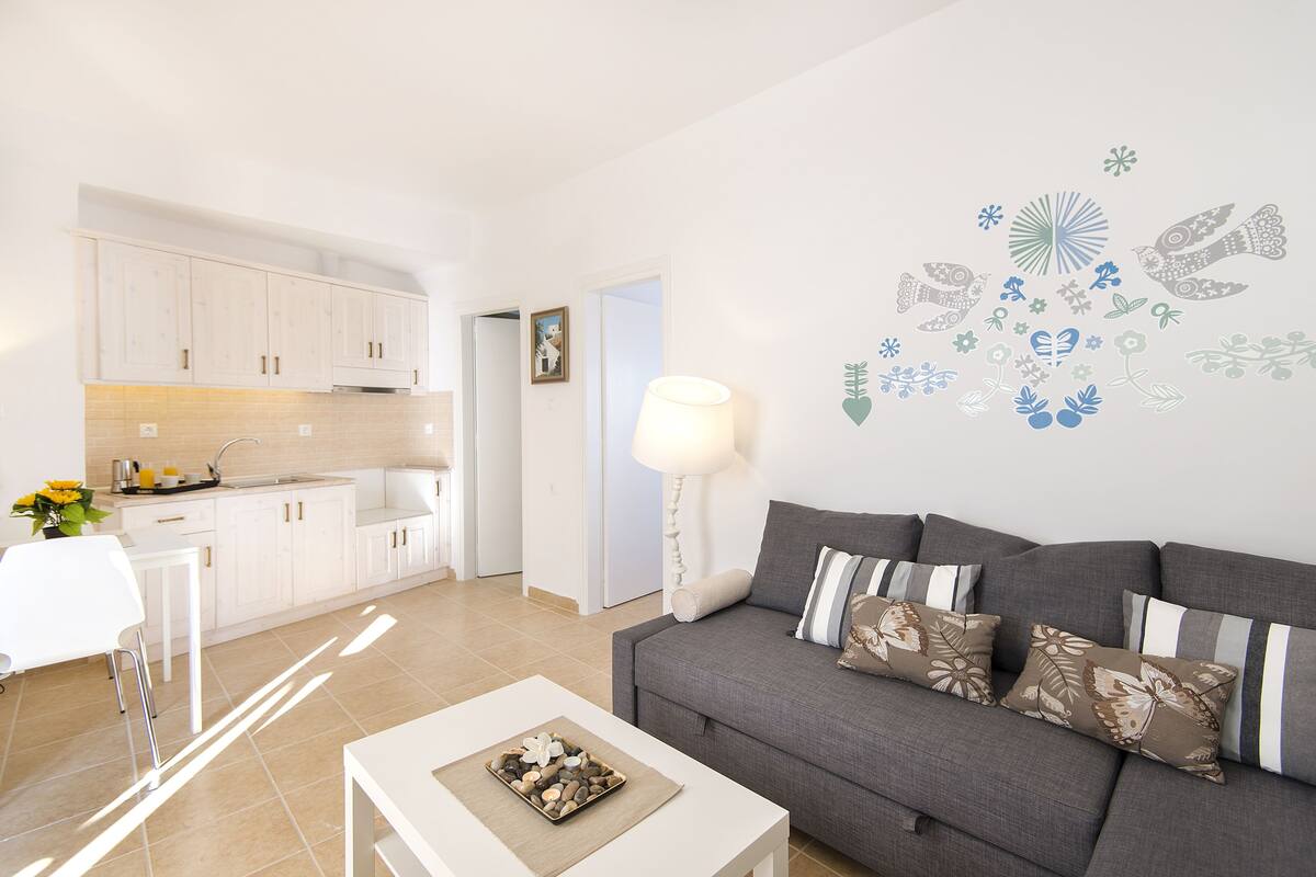 Una sala de estar moderna con un sofá gris, una mesa de centro blanca y una cocina con gabinetes blancos y un salpicadero de azulejos.