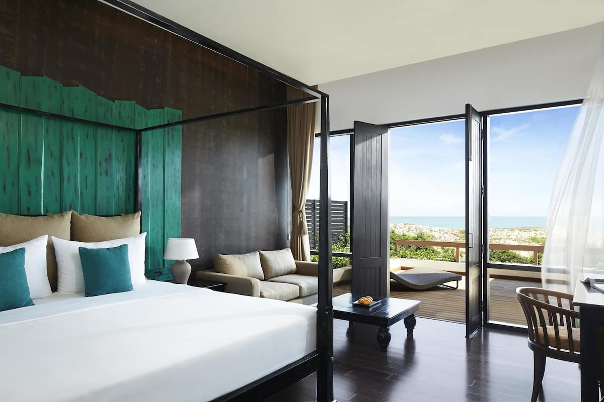 Habitación de hotel con una cama grande, un sofá, una mesa de comedor y un balcón con vistas a la playa.