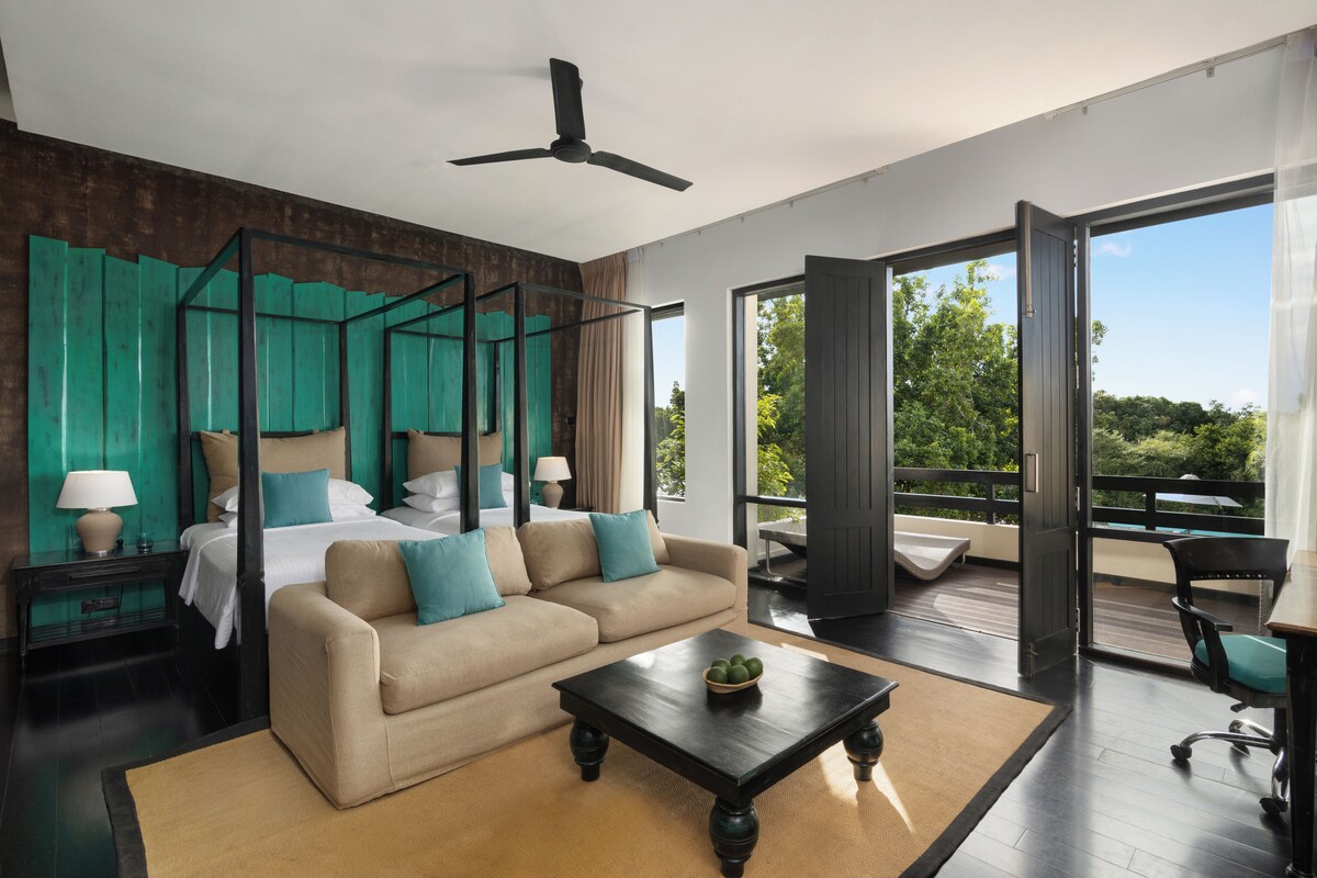 A modern bedroom with a four-poster bed, a sofa, a coffee table, and a balcony with a view of greenery.