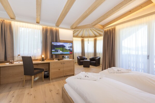 A hotel room with a wooden ceiling, a bed, a desk with a chair, a TV, and a seating area with chairs and a table. - Hotel Royal (Sesto)