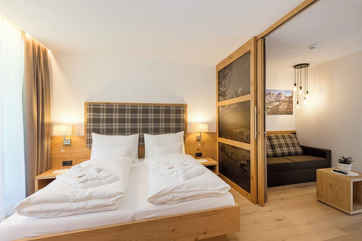 A modern hotel room with a wooden bed, two bedside tables with lamps, a dark sofa, and a small side table with a plant.