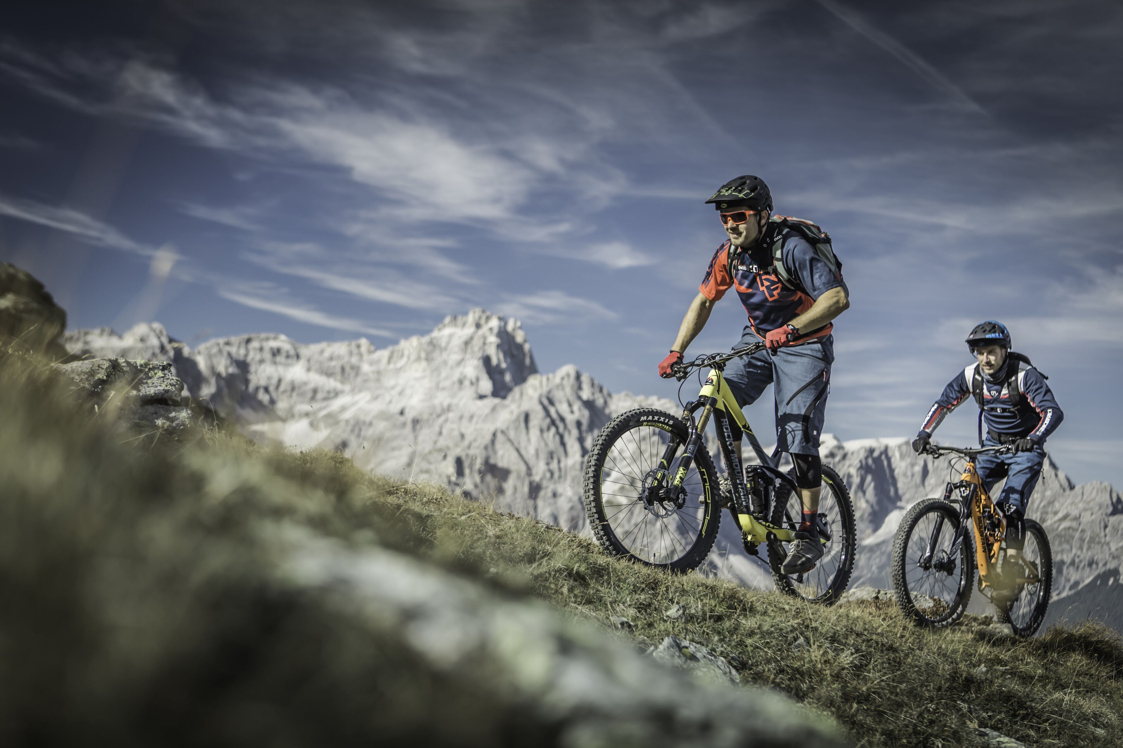 Two mountain bikers on a trail with rocky terrain and mountains in the background.