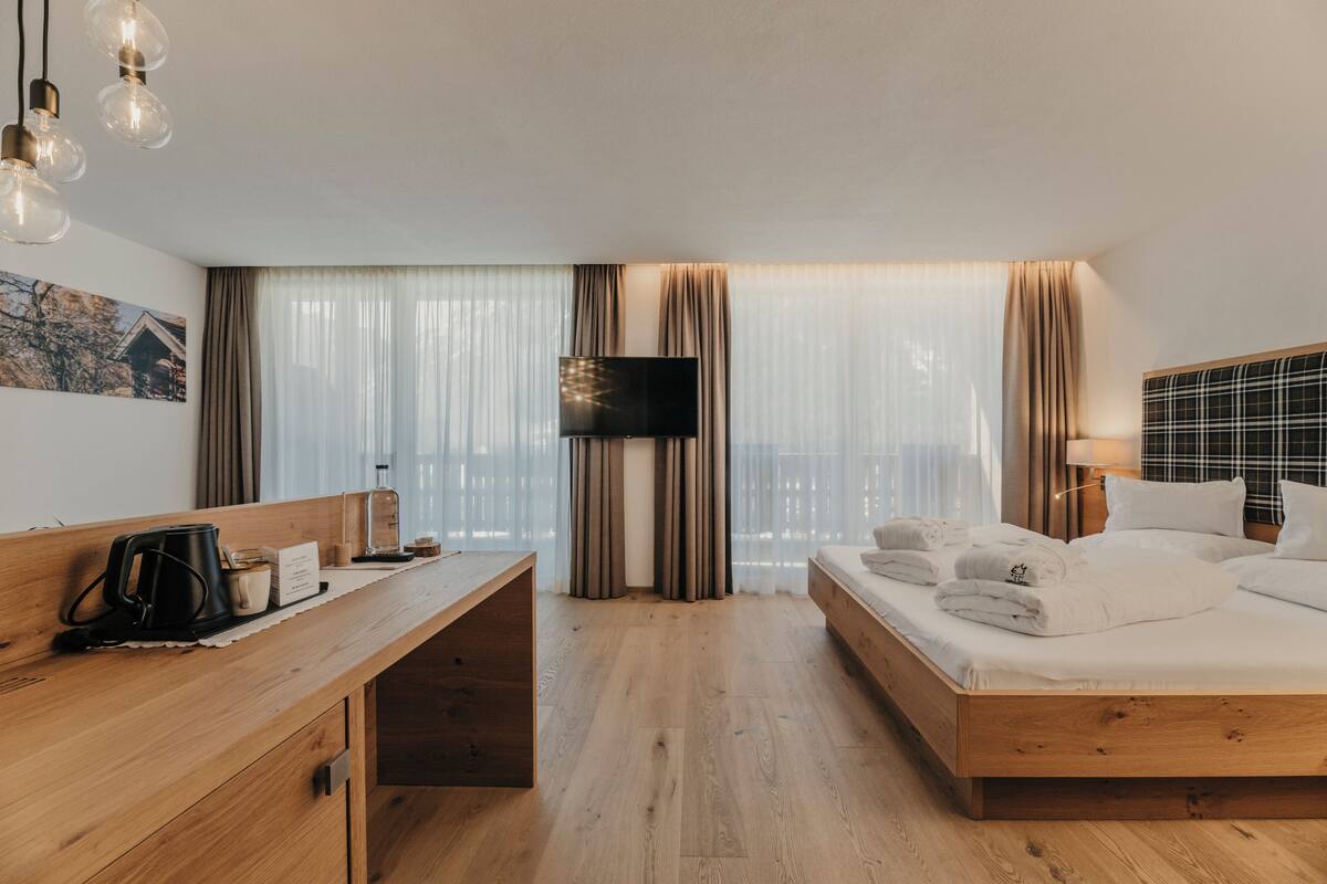 A hotel room with a wooden bed, a desk with a coffee maker, a flat-screen TV, and a large window with curtains.