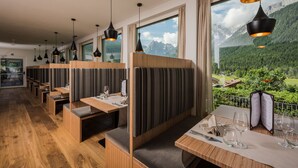 A restaurant with wooden booths and tables, large windows with mountain views, and pendant lighting. - Hotel Royal (Sesto)