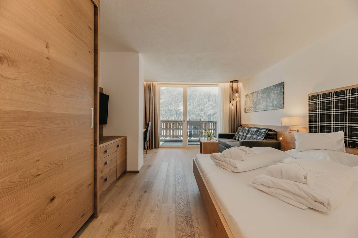 A modern hotel room with a wooden wardrobe, a bed with white linens, a sofa, a TV, and a balcony with a view of trees.
