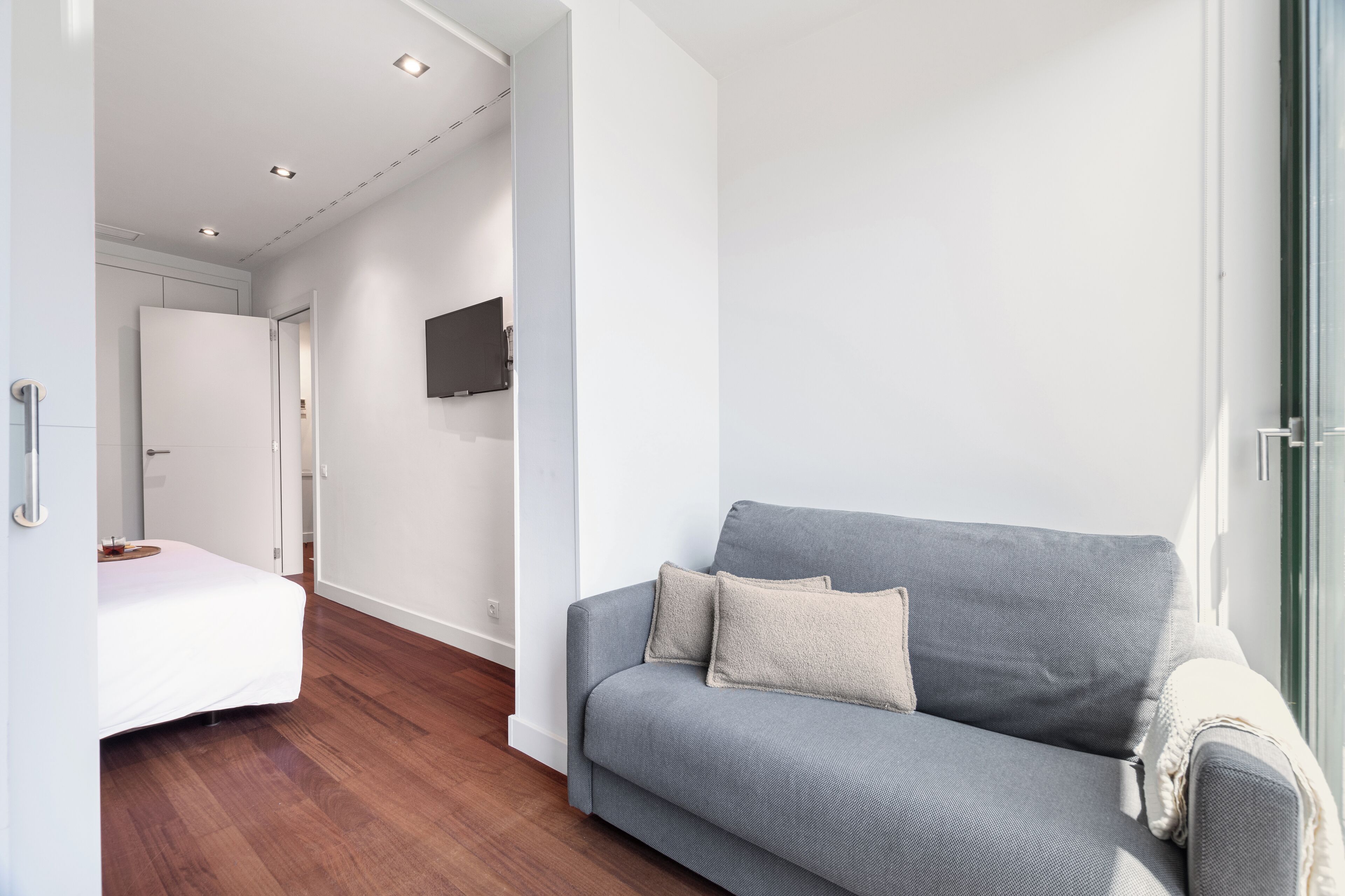 Een moderne woonkamer met een grijze bank, een wit bed en een flatscreen televisie aan de wand.