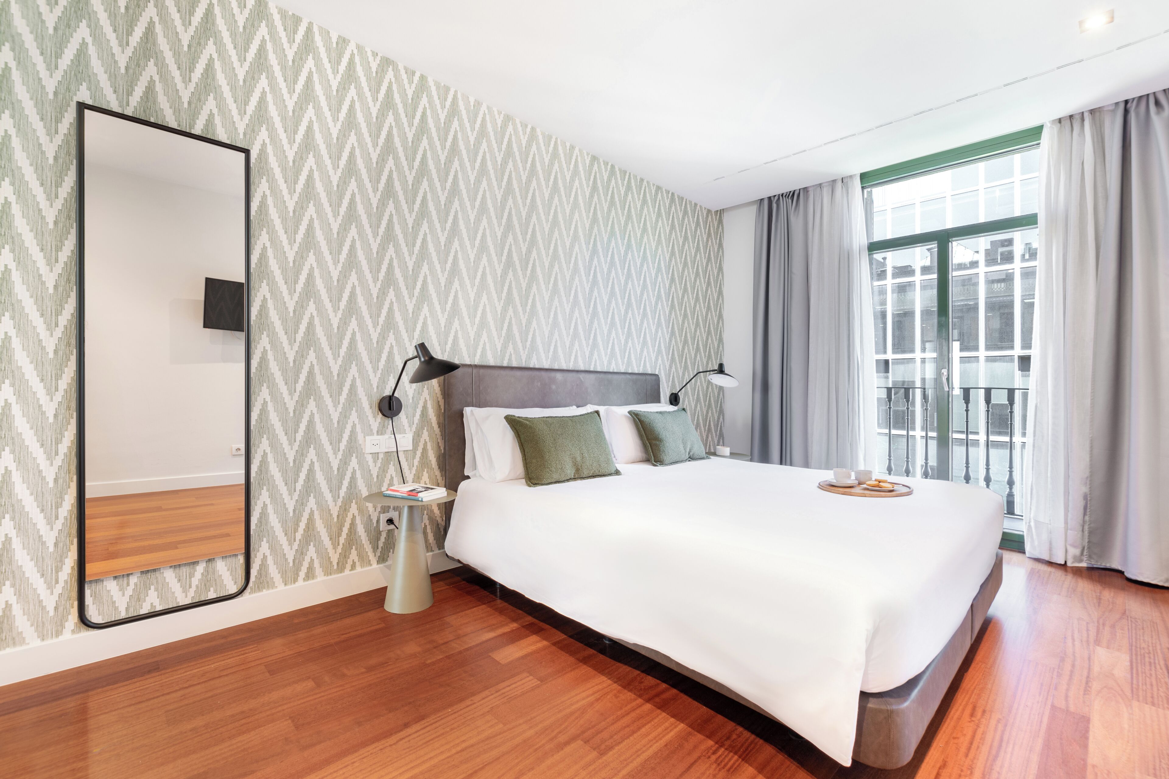 Ein modernes Hotelzimmer mit einem großen Bett, einem Nachttisch, einem Spiegel und Blick auf die Stadt durch große Fenster.
