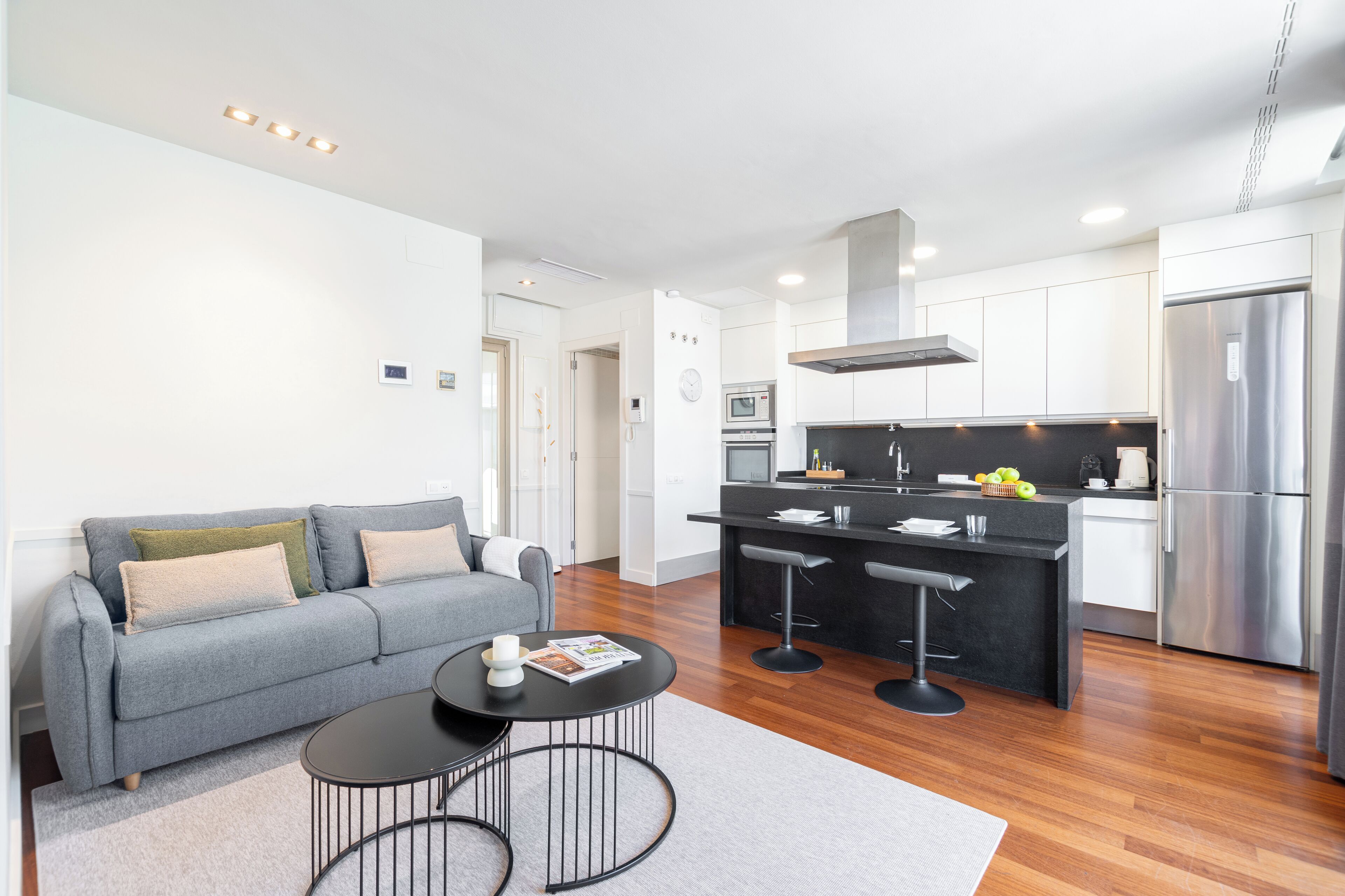 A modern living room with a grey sofa, a small round table, and a kitchen area with a black countertop, stainless steel appliances, and a bar stool.