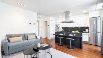 A modern living room with a grey sofa, a small round table, and a kitchen area with a black countertop, stainless steel appliances, and a bar stool.