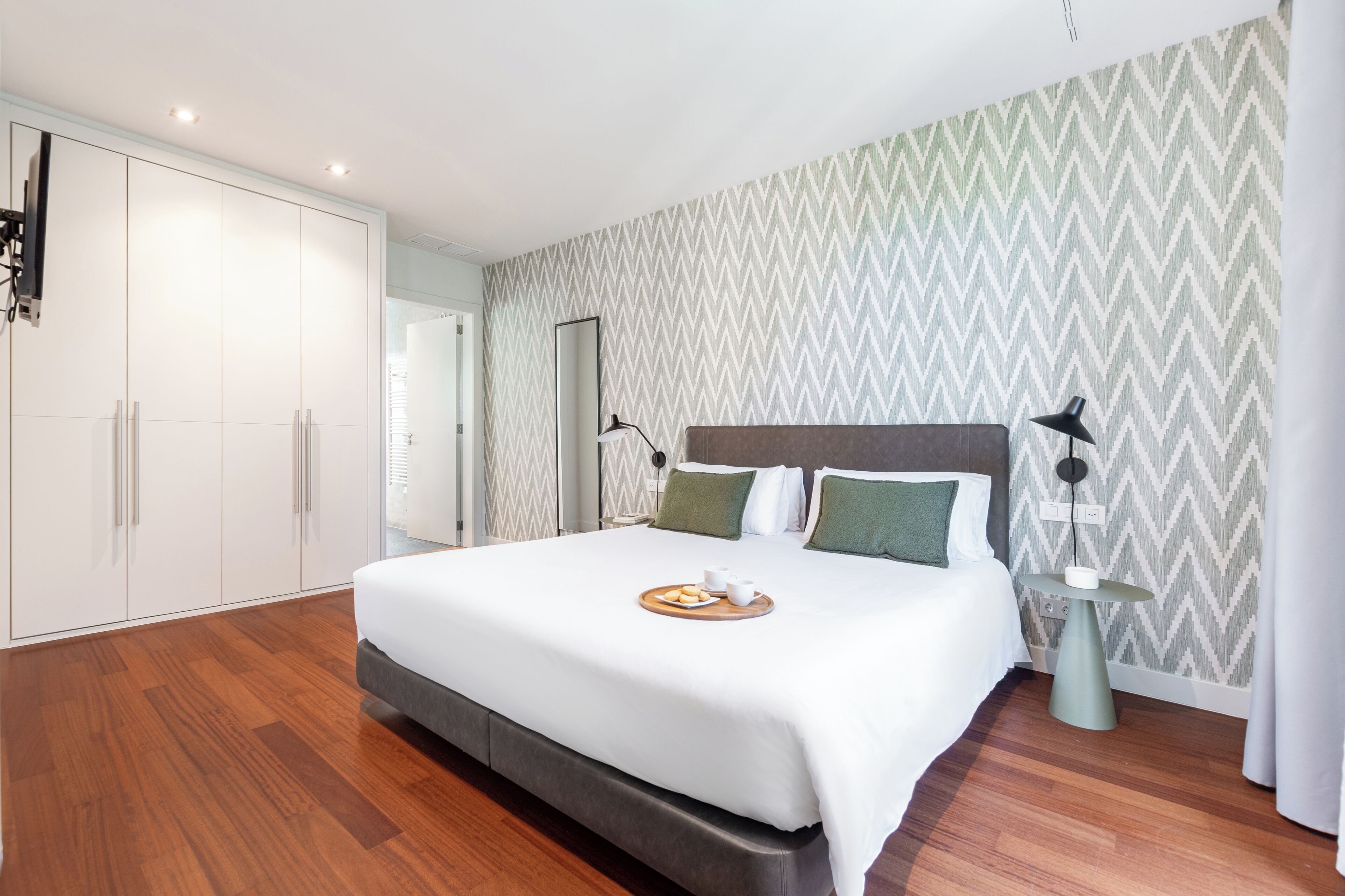Una habitación de hotel moderna con una cama grande, un armario, una mesita de noche y un espejo.