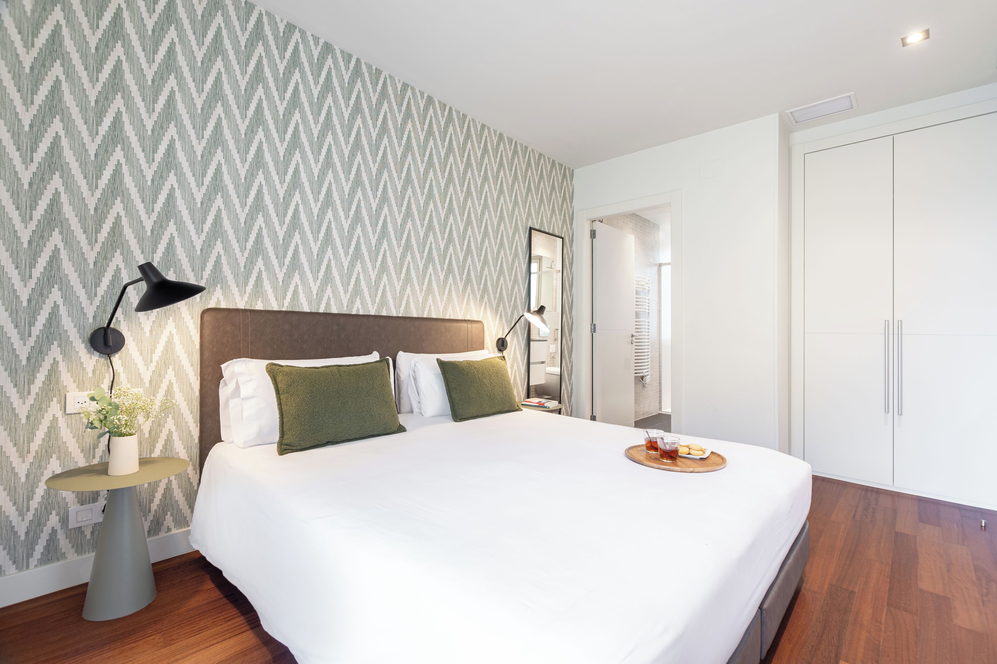 Habitación de hotel ordenada con una cama grande, una mesita de noche con lámpara, una pequeña mesa con planta y una bandeja con aperitivos.
