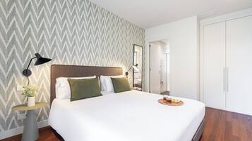 Habitación de hotel ordenada con una cama grande, una mesita de noche con lámpara, una pequeña mesa con planta y una bandeja con aperitivos.