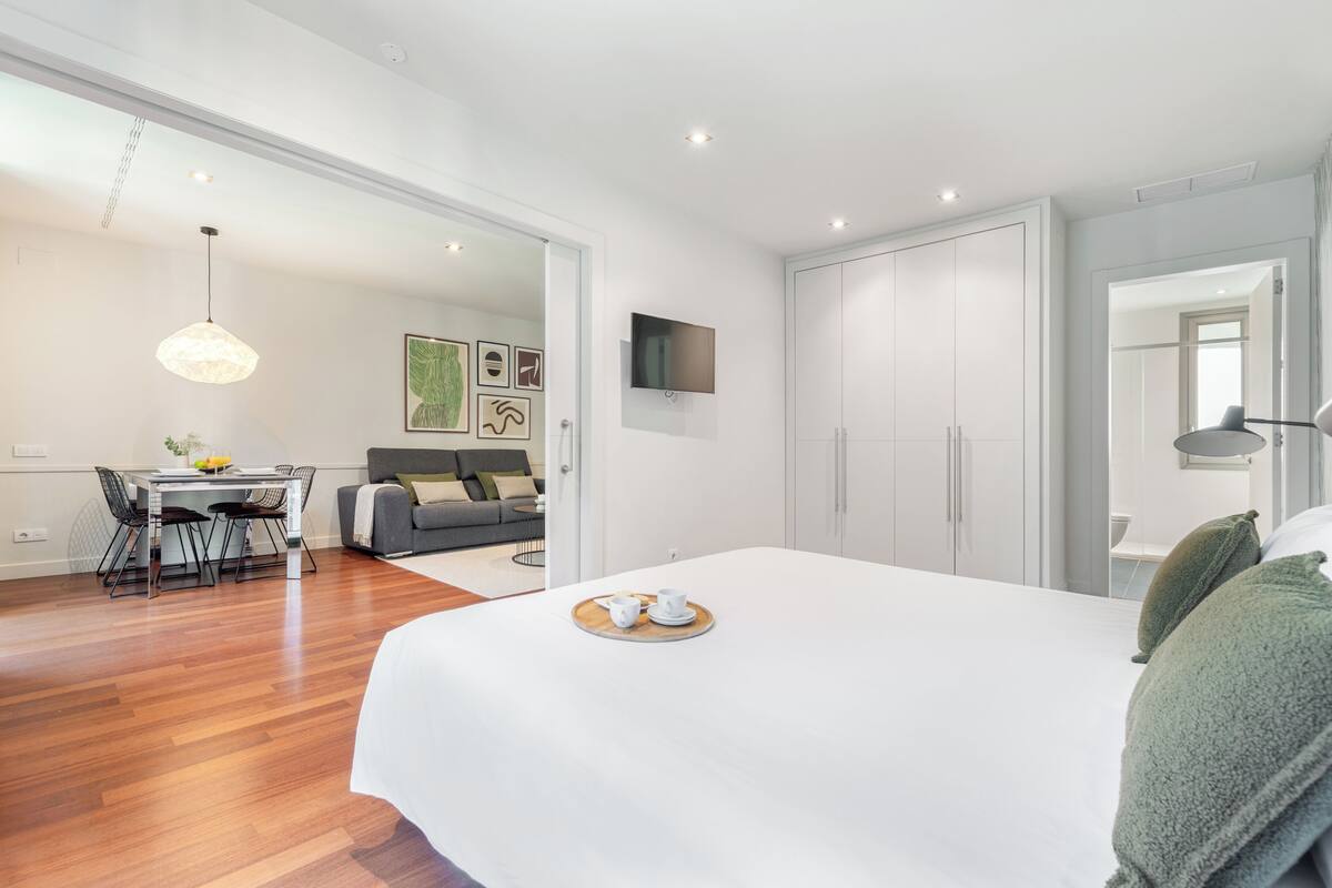 Une chambre moderne avec un grand lit, un coin repas comprenant une table et des chaises, un canapé gris et un téléviseur fixé au mur.