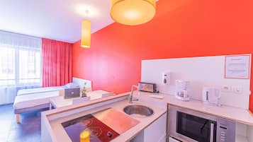 Una habitación de hotel moderna con una pared de acento roja, una kitchenette con microondas y tostadora, una cama con una computadora portátil y un ventanal con cortinas.