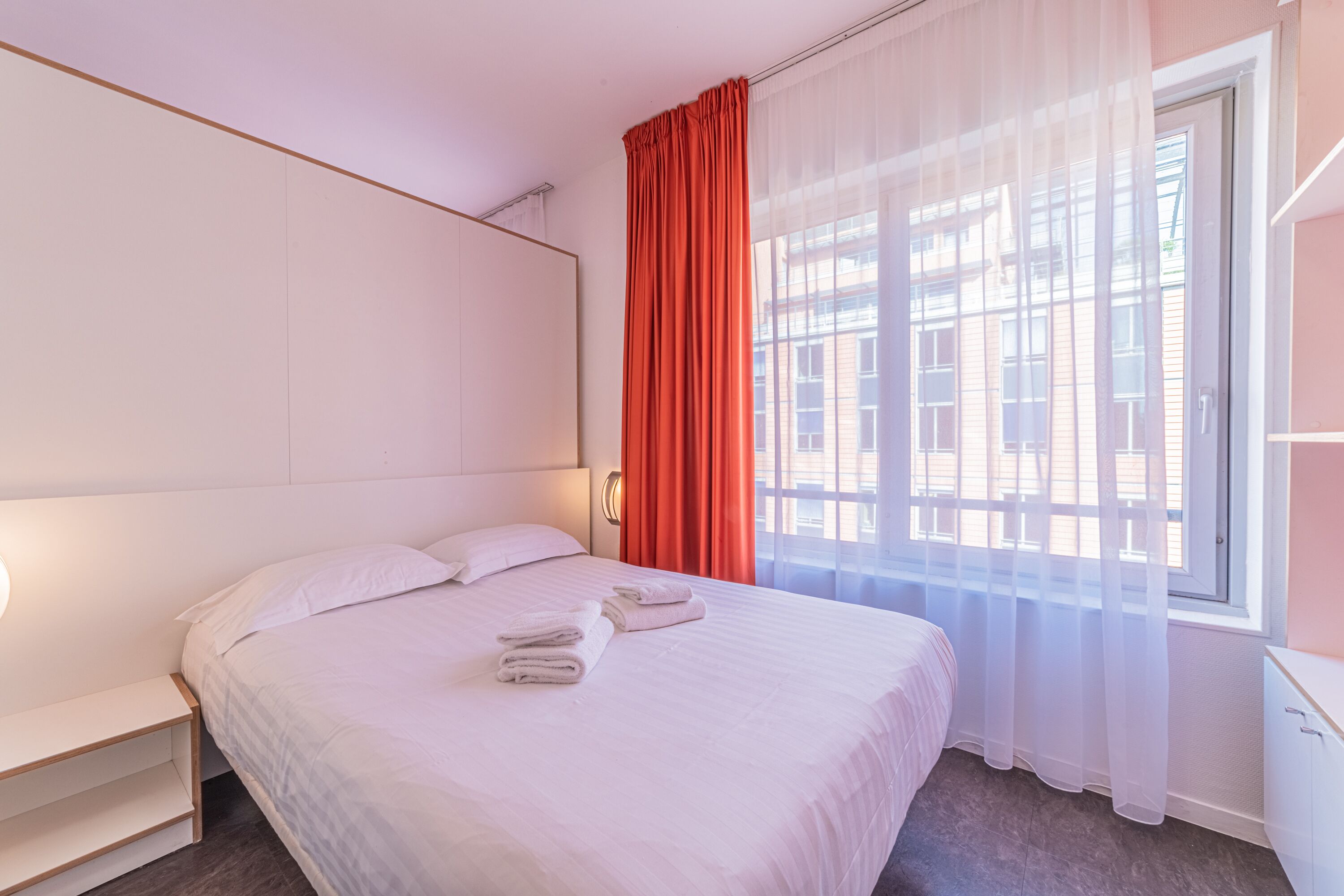 A hotel room with a large bed, a bedside table, a window with sheer curtains, and a view of buildings outside.