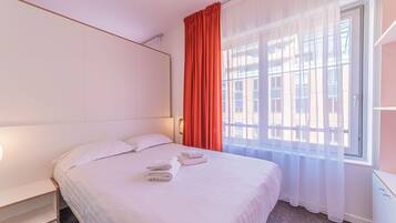 A hotel room with a large bed, a bedside table, a window with sheer curtains, and a view of buildings outside.